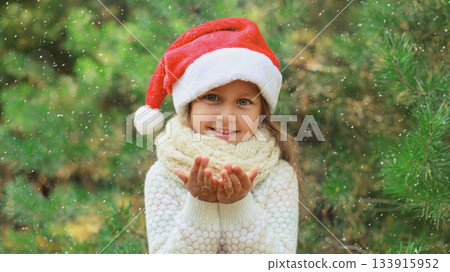 Christmas portrait of happy smiling little girl child in santa red hat blows snow in snowy forest 133915952