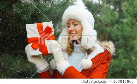 Happy smiling young woman holding red gift box on Christmas tree background in winter forest Happy smiling young woman holding red gift box on Christmas tree background in winter forest 133915963