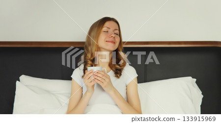 Happy smiling young woman enjoying with cup of hot coffee lying on bed at home Happy smiling young woman enjoying with cup of hot coffee lying on bed at home 133915969