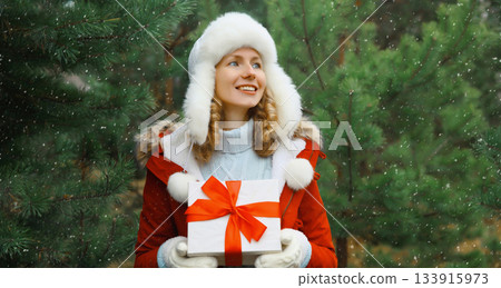 Happy smiling young woman holding red gift box on Christmas tree background in winter forest Happy smiling young woman holding red gift box on Christmas tree background in winter forest 133915973