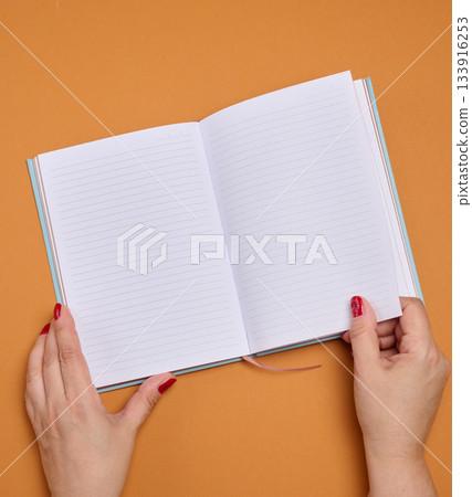 Female hands holding an open notebook with clean, lined pages on an orange background. 133916253