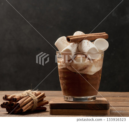 A glass of hot chocolate or cocoa with lots of cinnamon-dusted marshmallows on a wooden background. 133916323