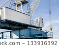 Industrial crane structure rises under clear sky, showcasing metal framework, cabin, pulley and hook. Scene highlights machinery, steel geometry and port atmosphere with strong sunlight and shadows 133916392