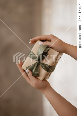 Close up of hands holding beige gift box with green ribbon against blurred festive lights 133916407