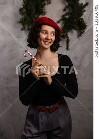 Caucasian woman holding candy cane in festive room with grey wall and green garland 133916409
