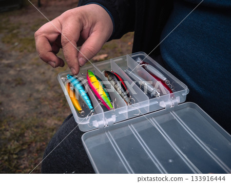Hand-picking brightly colored lures. The tackle box is open on the lap, and the angler is selecting lures and plastic compartments with bright trim. Get the rods ready. Hand-picking brightly colored lures. The tackle box is open on the lap, and the angler is selecting lures and plastic compartments with bright trim. Get the rods ready. 133916444