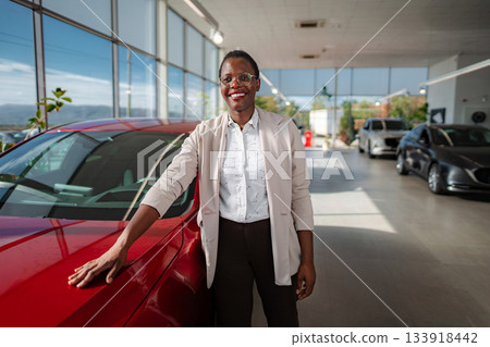 Smiling woman standing next to new red car in showroom 133918442