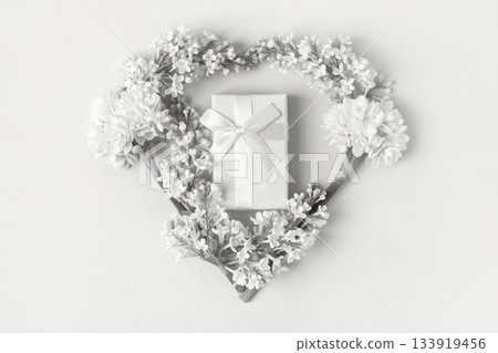 Demonstrating the colors of 2026 - Cloud Dancer. Romantic composition in the shape of a heart of white lilac flowers, white daffodils and gift box with satin bow on gray background. 133919456