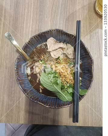Beef noodle soup bowl, basil garnish, fried garlic, chopsticks, spoon, rustic ceramic, wooden table Beef noodle soup bowl, basil garnish, fried garlic, chopsticks, spoon, rustic ceramic, wooden table 133920530
