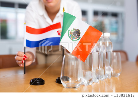A woman in a white blouse with friendly smile arrangs flags of Mexico and Netherlands for meeting. 133921612