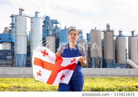Sad young woman worker with flag of Georgia against background of factory Sad young woman worker with flag of Georgia against background of factory 133921635
