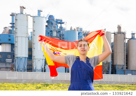 Sad young woman worker with flag of Spain against background of factory 133921642