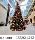 Tall Christmas tree in an indoor atrium, decorated with red, gold, and white ornaments. The building features a high glass ceiling with metal beams and brick walls with arched openings 133921842