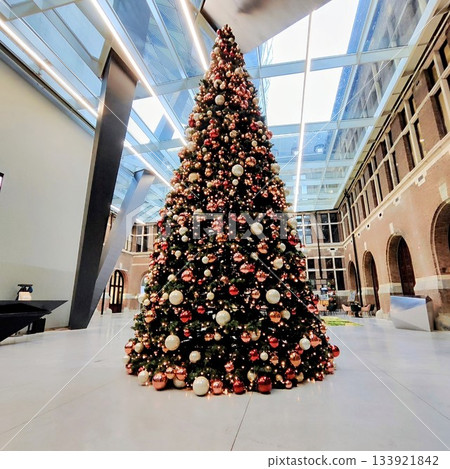 Tall Christmas tree in an indoor atrium, decorated with red, gold, and white ornaments. The building features a high glass ceiling with metal beams and brick walls with arched openings Tall Christmas tree in an indoor atrium, decorated with red, gold, and white ornaments. The building features a high glass ceiling with metal beams and brick walls with arched openings 133921842