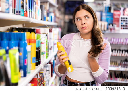 Thoughtful woman standing near shelves of hair care products and puzzled looking at her hair during choosing hairspray Thoughtful woman standing near shelves of hair care products and puzzled looking at her hair during choosing hairspray 133921843