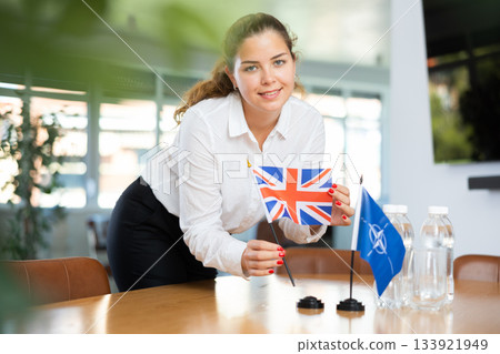 Woman secretary prepares an office for negotiations - she places flags of NATO (OTAN) and Great Britain table Woman secretary prepares an office for negotiations - she places flags of NATO (OTAN) and Great Britain table 133921949