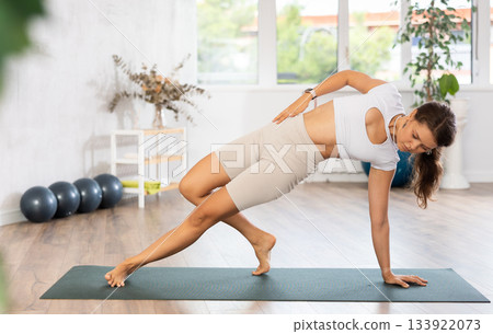 Young woman yoga trainer performing side plank pose on black rug 133922073