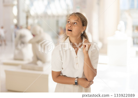 Girl visitor looks with interest at the sculpture in the museum hall, holding an information booklet in her hands 133922089