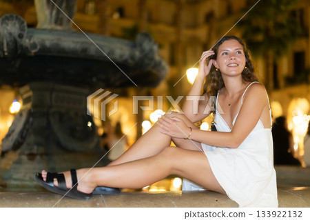 Night Royal square in Barcelona, girl in sundress is sitting on fountain fencing. 133922132