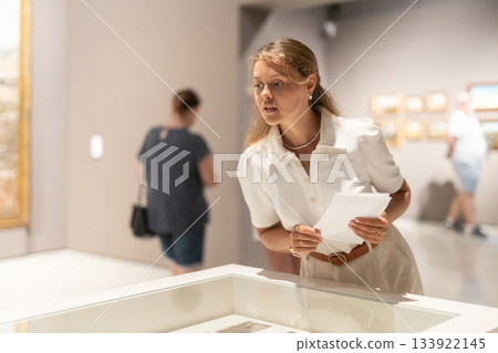 Young female visitor stands near glass display case and examines valuable exhibit Young female visitor stands near glass display case and examines valuable exhibit 133922145