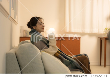 一位年輕女子在夜晚燈光明亮的房間裡喝著熱飲。冬日里女子的形象。溫暖的白熾燈光映照著她。 一位年輕女子在夜晚燈光明亮的房間裡喝著熱飲。冬日里女子的形象。溫暖的白熾燈光映照著她。 133922161