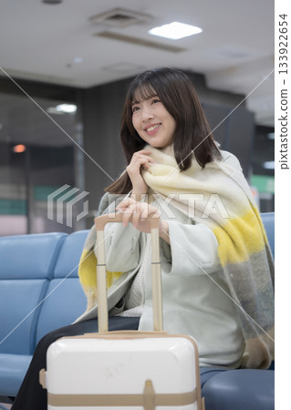 Travel scene of a young woman with a suitcase in the airport lobby in winter, sitting in the lobby waiting for boarding Travel scene of a young woman with a suitcase in the airport lobby in winter, sitting in the lobby waiting for boarding 133922654