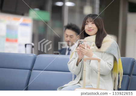 冬日機場大廳裡，一位年輕女子拖著行李箱，坐在候機廳等待登機。 133922655