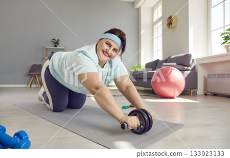 Happy, overweight woman doing fitness exercise with ab roller during her sports workout at home 133923133