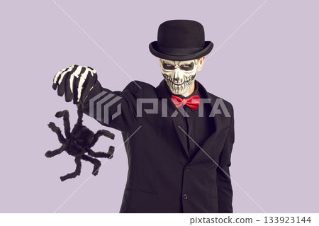 Scary mad man in halloween costume with black spider in hand looking at camera at purple background. Scary mad man in halloween costume with black spider in hand looking at camera at purple background. 133923144
