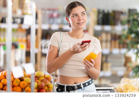 Young woman customer at shop scans QR code on yellow smoothie bottle using phone camera 133924259