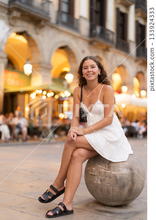 Evening Royal square in Barcelona, girl in sundress is sitting on stone ball 133924333