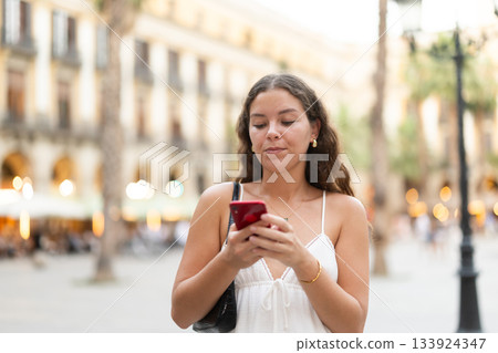 In evening, smiling girl with smartphone walks along city street Placa Real 133924347