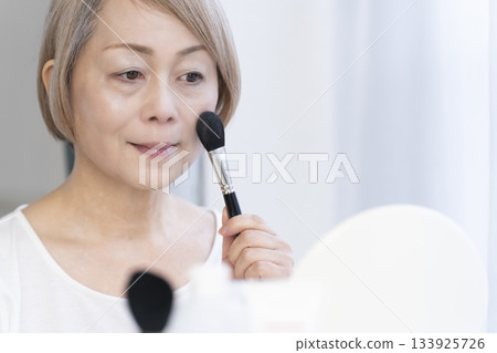 Senior woman putting on makeup by a bright window Senior woman putting on makeup by a bright window 133925726