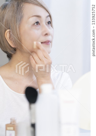 Senior woman putting on makeup by a bright window Senior woman putting on makeup by a bright window 133925752