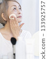 一位老婦人在明亮的窗邊化妝。 133925757