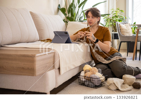 Feminine hipster man crochet handmade use hook knitting needles and wool yarn relaxing on couch Feminine hipster man crochet handmade use hook knitting needles and wool yarn relaxing on couch 133926821