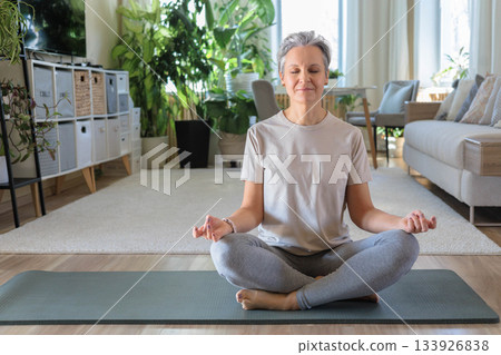 Smiling mature gray haired sport woman sits on yoga mat in lotus position. elderly lady meditates Smiling mature gray haired sport woman sits on yoga mat in lotus position. elderly lady meditates 133926838