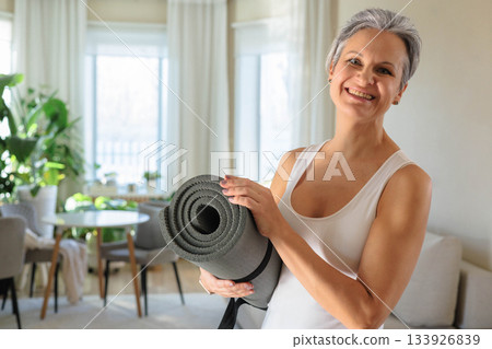 Beautiful gray-haired mature middle-aged woman holds a rolled-up yoga mat at home. Beautiful gray-haired mature middle-aged woman holds a rolled-up yoga mat at home. 133926839