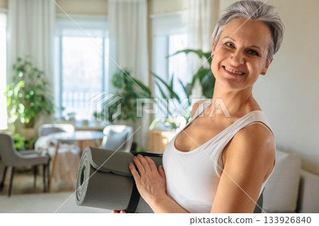 An elderly mature woman with gray hair is smiling and holding a folded yoga mat. An elderly mature woman with gray hair is smiling and holding a folded yoga mat. 133926840