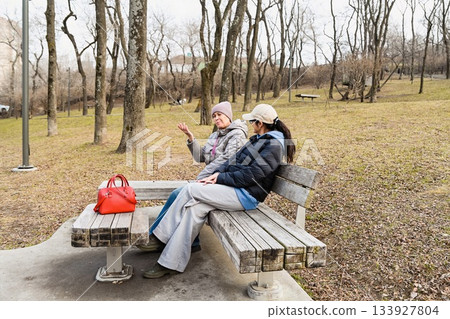 Two Russian friends aged 50 and 60 sit in Nagorny Park Vladivostok, enjoying calm spring conversation that supports emotional balance, connection and wellbeing. 133927804