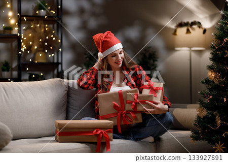 A woman wearing a Santa hat smiles while holding wrapped Christmas presents on a couch A woman wearing a Santa hat smiles while holding wrapped Christmas presents on a couch 133927891