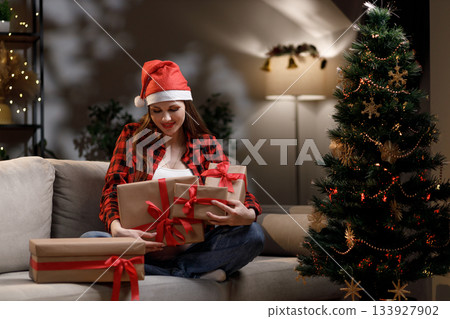 A woman wearing a Santa hat is holding wrapped Christmas presents near a decorated tree A woman wearing a Santa hat is holding wrapped Christmas presents near a decorated tree 133927902