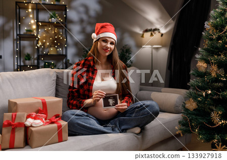 A pregnant woman wearing a Santa hat holds an ultrasound image while sitting on a couch near a Christmas tree A pregnant woman wearing a Santa hat holds an ultrasound image while sitting on a couch near a Christmas tree 133927918