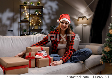 A pregnant woman wearing a Santa hat sits on a couch with wrapped gifts, celebrating the holiday season 133927921