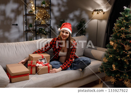 A pregnant woman wearing a Santa hat smiles while sitting on a couch near a Christmas tree and wrapped gifts 133927922