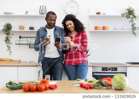 A diverse couple smiles while looking at their phones in a modern kitchen, with food on the counter. 133929026