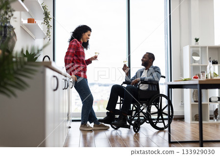 A diverse couple enjoys a celebratory moment in a bright, modern kitchen, with one person in a wheelchair. 133929030