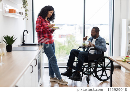 A diverse couple enjoys a healthy salad in a bright, modern kitchen. One person is in a wheelchair. 133929048