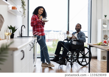 A diverse couple enjoys a meal together in a modern kitchen, with one person using a wheelchair. 133929049