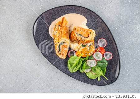 Crispy salmon wellington with mashed potato, cream sauce and fresh spinach salad served on a modern dark plate 133931103